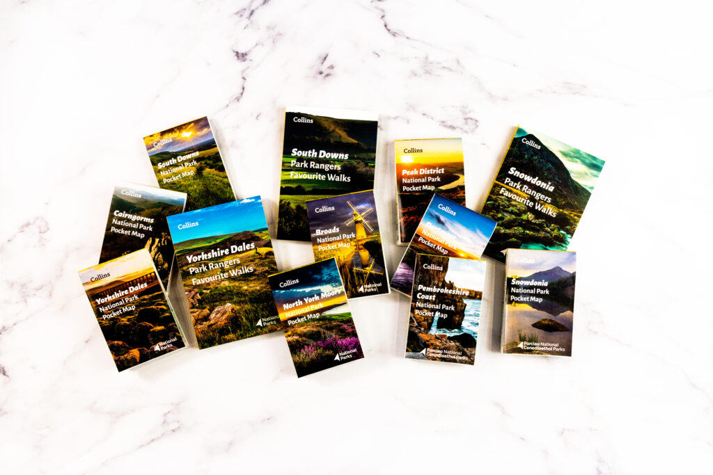 Walking guidebooks and pocket maps for UK national parks and trails spread out on white marble.