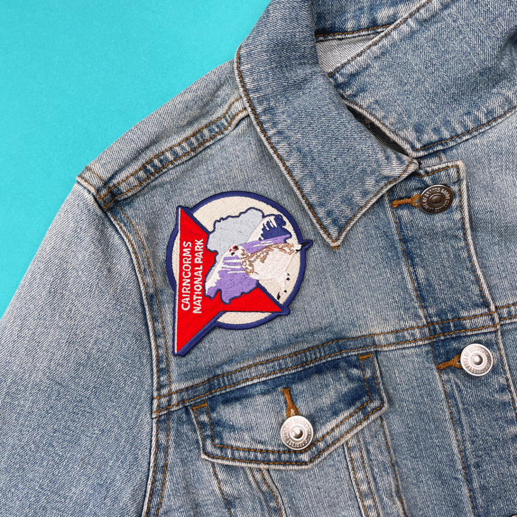 Denim jacket with a colorful Crater Lake patch on chest pocket, shown on a blue background.