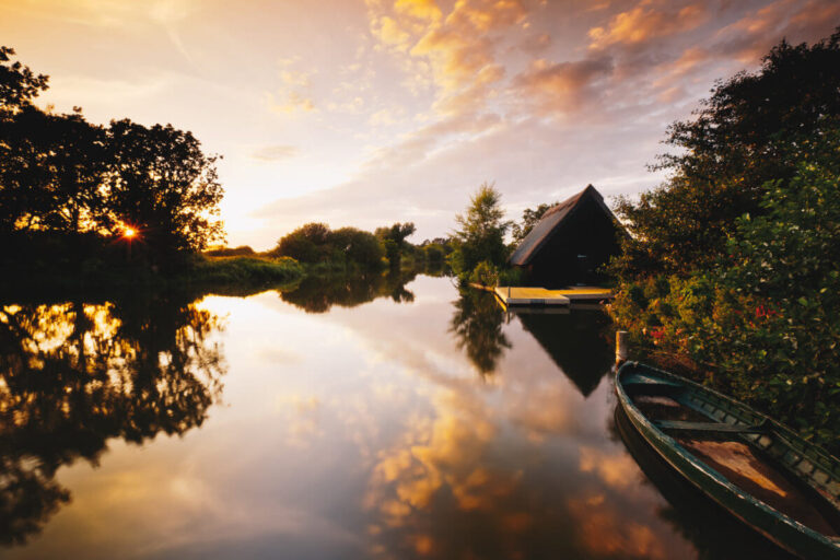 Lakeside at sunset: cabin, trees, boat; calm waters reflect orange and purple hues.