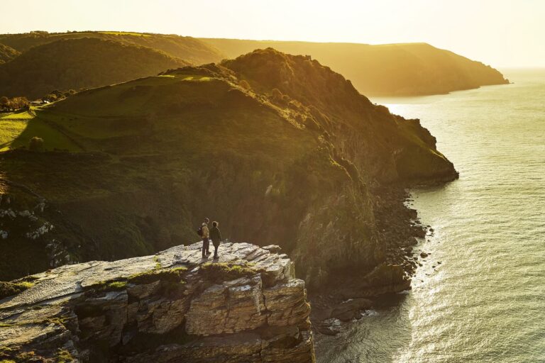 Two hikers atop valley of rocks exmoor, overlooking water; sunset casts a golden glow over serene landscape.