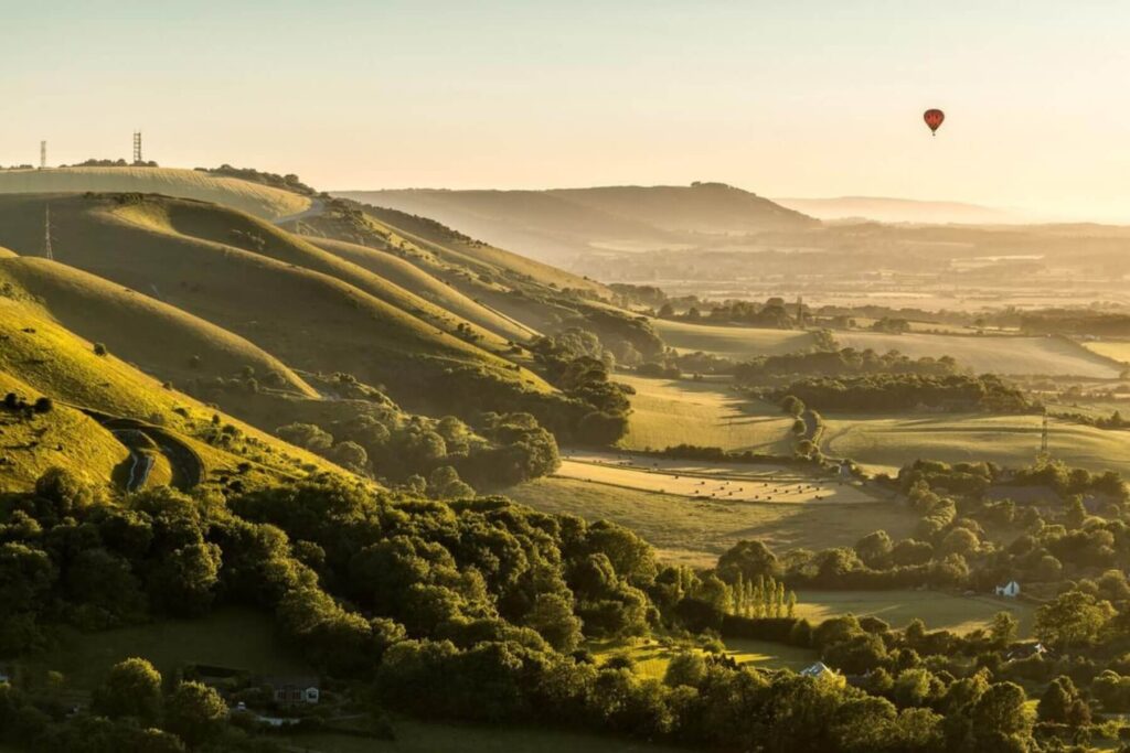 Rolling green hills, clear sky; sunlit with shadows. A red hot air balloon floats distantly.