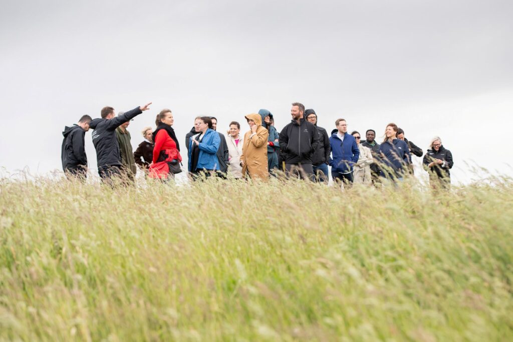 People in jackets stand in a grassy field under clouds; one points while others converse.