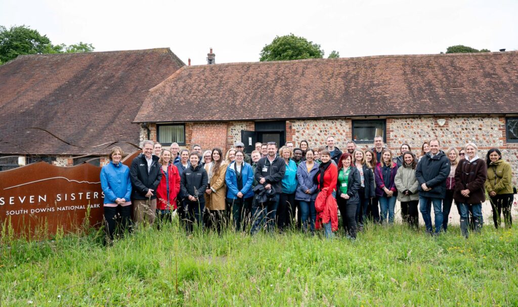 A group smiles by a brick building and South Downs sign, surrounded by grass and wildflowers.
