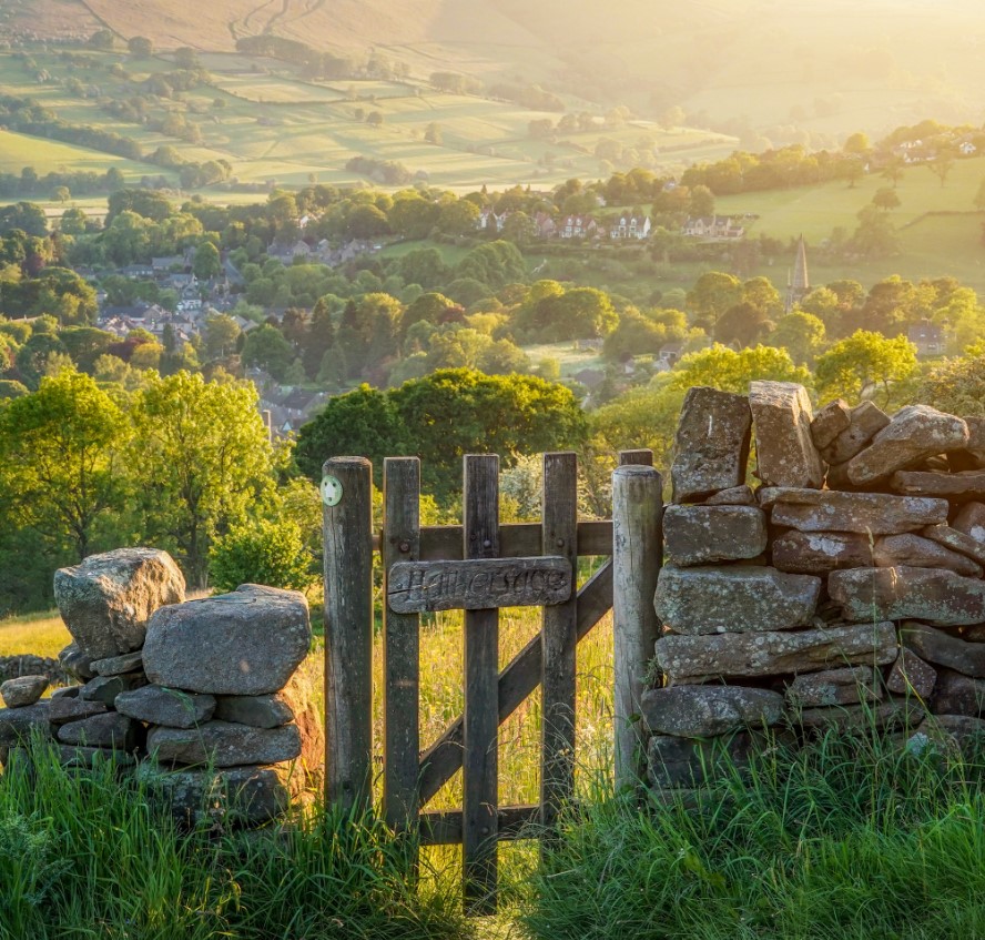 A wooden gate in a stone wall overlooks green fields, trees, and a distant village in rolling hills.