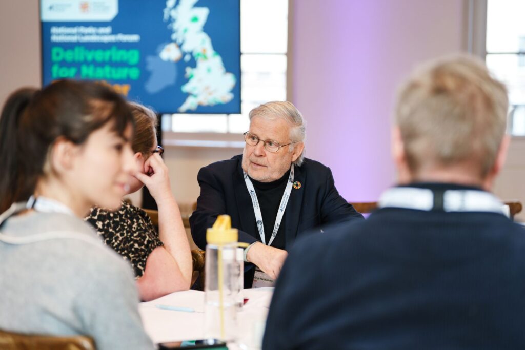 People sit round a table at a conference; “Delivering for Nature” appears on a screen with UK map.