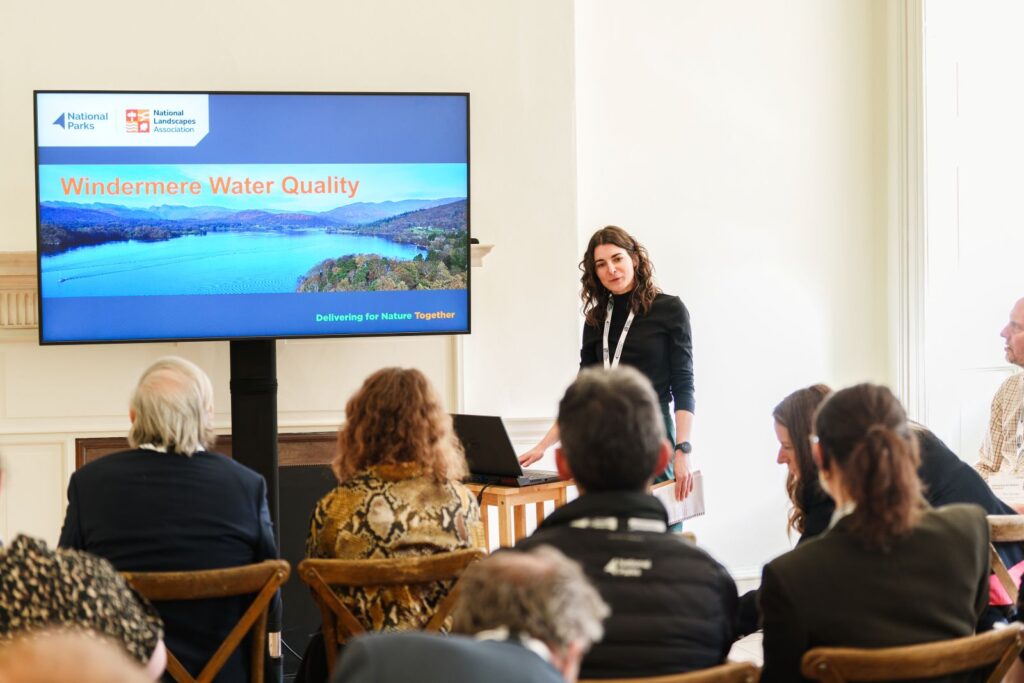A woman presents Windermere Water Quality beside a screen to an audience in a bright room.