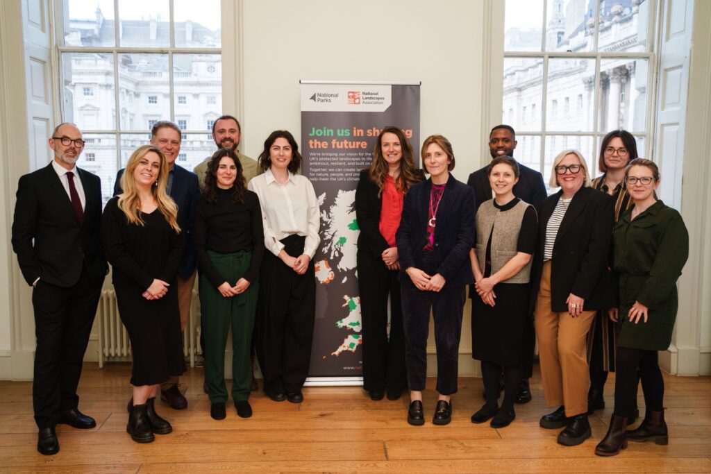 Thirteen people smile in front of a "Join us in shaping the future" banner in a bright, airy room.