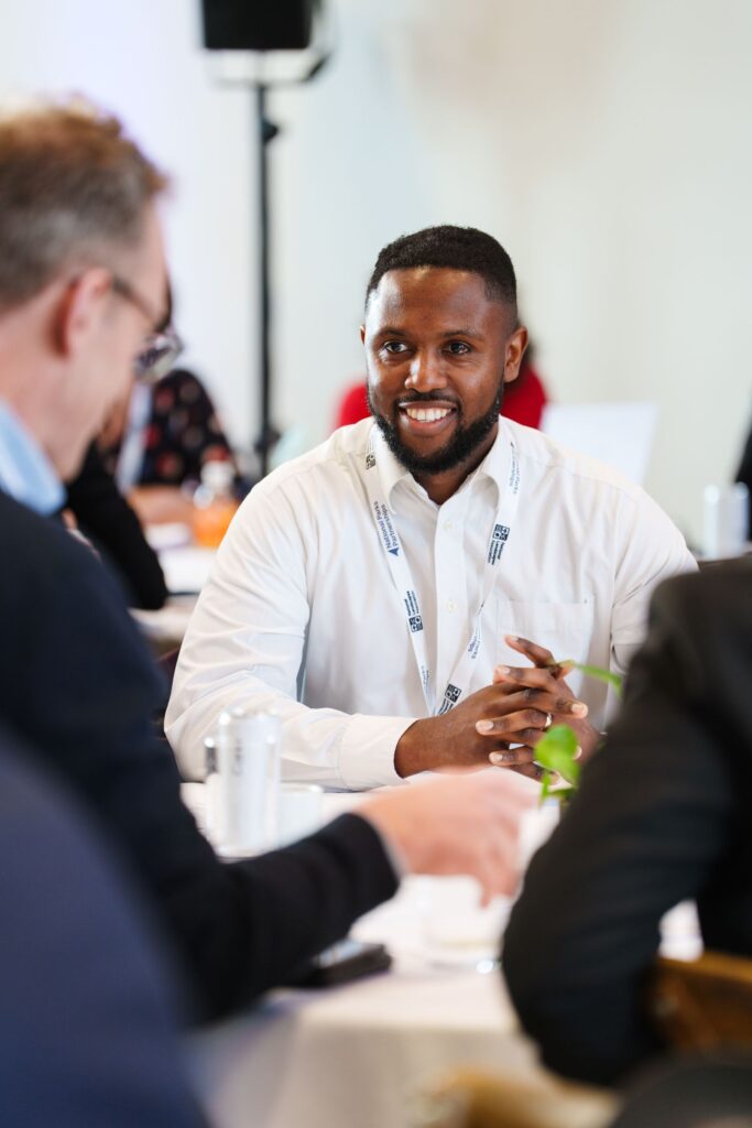 Man in white shirt at a table, smiling and talking with others during a meeting, wearing a lanyard.