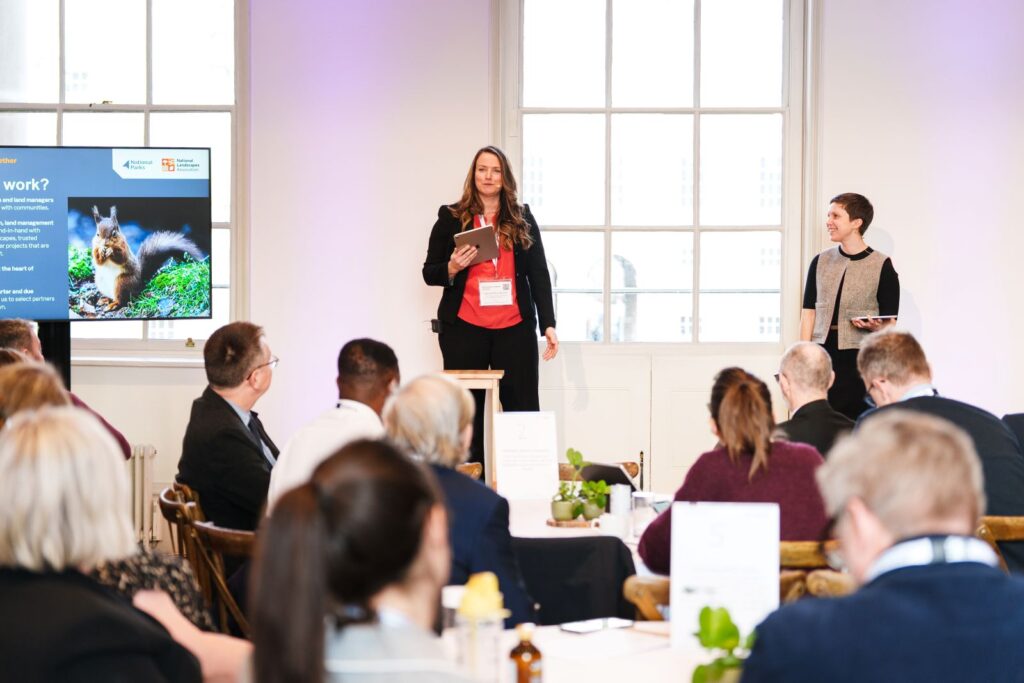 Woman stands on chair with tablet addressing audience; another person, attendees, slide in background.