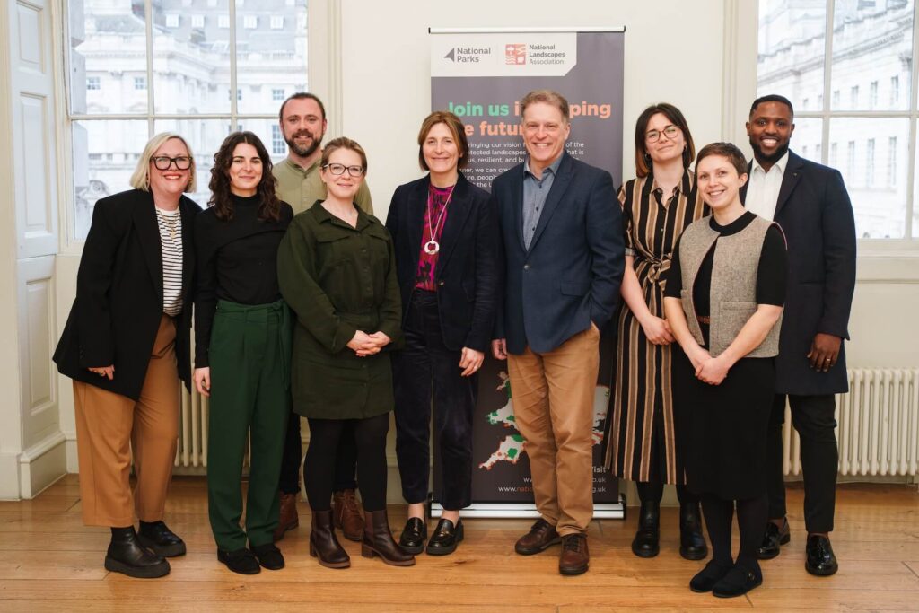 National Parks Partnerships Team Members - Nine people stand smiling indoors by a National Parks banner, casually dressed with windows behind them.